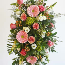 Tall standing spray of pink gerbera daisies and roses with white blooms and green accents on an easel.