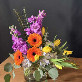 Bright mixed bouquet with orange gerbera daisies, purple flowers, and yellow tulips in a glass vase