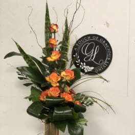 Tall orange rose arrangement in a gold vase with dark foliage