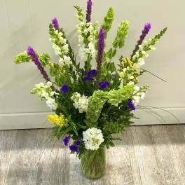 Tall mixed flower arrangement in a glass vase with purple, white, yellow, and green blooms