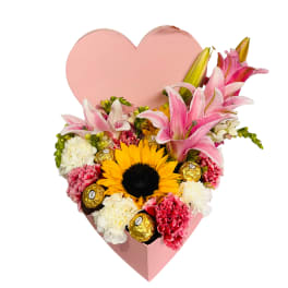 Heart-shaped pink box with pink lilies, carnations, a sunflower, and gold-wrapped chocolates