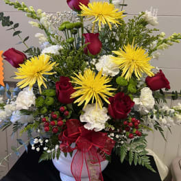 Red roses and yellow spider mums arranged in a white vase with a red ribbon.
