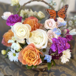 Basket arrangement of roses, daisies, and purple blooms with a butterfly accent