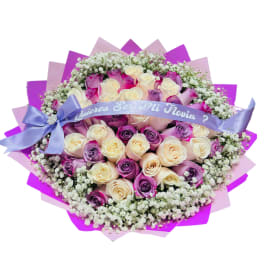Round bouquet of cream and lavender roses with baby’s breath, wrapped in pink paper and a lavender ribbon.
