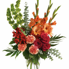 Tall mixed arrangement of orange blooms, red roses, and peach carnations in a clear glass vase