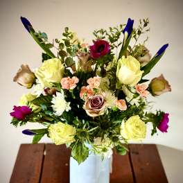 Mixed bouquet of yellow roses, peach carnations, and purple blooms in a white vase on a wooden table