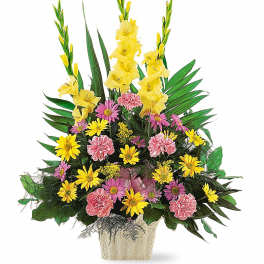 Tall arrangement of yellow gladiolus, pink carnations, and yellow daisies with lush greenery in a white container