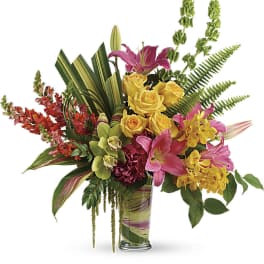 Tall tropical arrangement with pink lilies, yellow roses, orchids, and tropical foliage in a clear glass vase