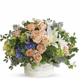 Low arrangement of peach roses, white lilies, and blue and green hydrangeas in a white ceramic container