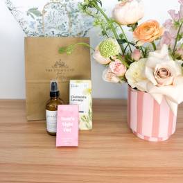 Pastel flower arrangement in a pink striped vase with gift items beside it