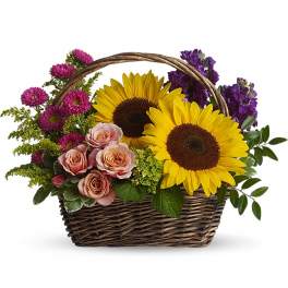 Woven basket of yellow sunflowers, pink roses, and purple flowers with a handle.