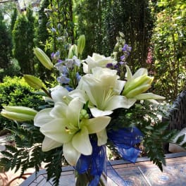 White lily and rose arrangement with blue ribbon and purple accents in a clear vase on an outdoor table