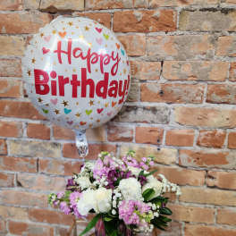 Birthday balloon and small pink and white flower arrangement in a clear vase on a stand.