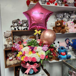 Mixed flower arrangement in a black hat box with pink balloons and teddy bears behind it