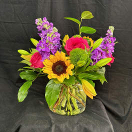 Mixed bouquet in a clear glass vase with sunflowers, pink roses, and purple blooms