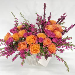 Orange and magenta roses in a white vase with purple filler flowers