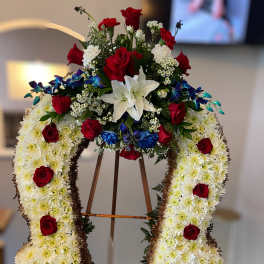 Horseshoe-shaped standing spray of white mums with red roses, lilies, and blue floral accents on an easel.