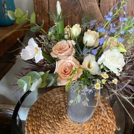 Peach roses, white blooms, and purple delphinium in a ribbed gray vase on a woven mat.