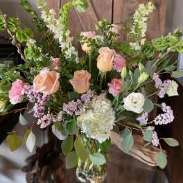 Tall arrangement of peach roses, white hydrangea, pink flowers and green bells of Ireland in a glass vase