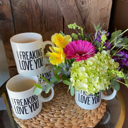 White 'I freaking love you' mugs, one filled with pink, yellow, and green flowers on a woven mat.