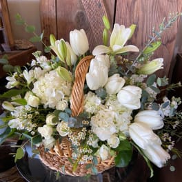 White tulips, lilies, and hydrangeas arranged in a woven basket against a rustic wooden backdrop