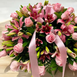 Low arrangement of pink roses and pink lilies with buds, accented by a large pink ribbon bow.