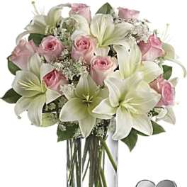Pink roses and white lilies in a clear glass vase with a heart keepsake