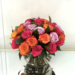 Round bouquet of hot pink, orange, and lavender roses arranged in a clear glass vase.