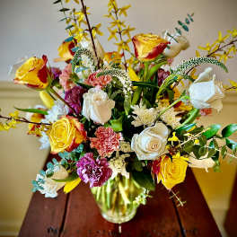 Mixed arrangement of yellow and white roses with pink carnations in a clear glass vase on a wooden table.