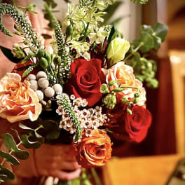 Hand-tied bouquet with red and orange roses, peach blooms, and small white accent flowers.