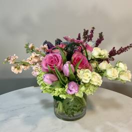 Low glass vase of pink roses, purple tulips, cream spray roses and mixed blooms.