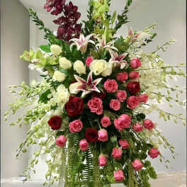 Tall standing floral spray with red and pink roses, lilies, orchids and trailing greenery.