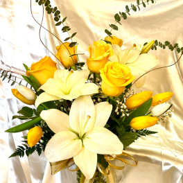 Yellow roses and white lilies arranged in a glass vase with a gold ribbon.
