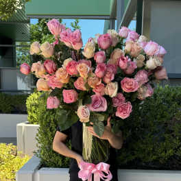 Large bouquet of pink and peach roses in a tall glass vase with ribbon, held by a person outdoors