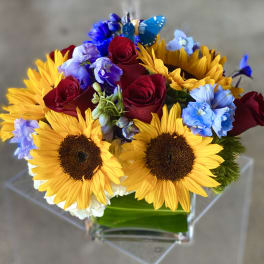 Bouquet of sunflowers, red roses, and blue flowers in a clear vase