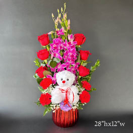 Red roses and pink orchids arranged with a white teddy bear in a red vase.