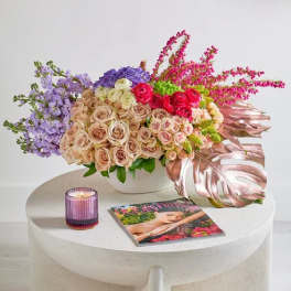 Mixed bouquet of roses and purple flowers in a white vase with a candle and magazine