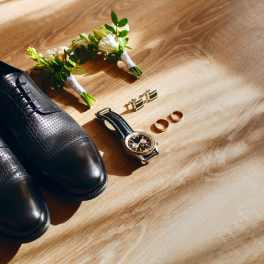 Black dress shoes with boutonniere, cufflinks, rings, and watch on wood floor