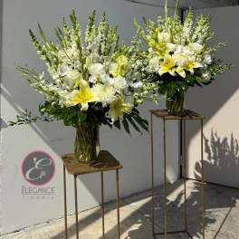 Two tall white and yellow floral arrangements in glass vases on stands