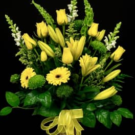 Tall yellow and green floral arrangement with lilies, roses, and daisies in a patterned cylinder vase with bow