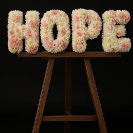 Floral letters spelling HOPE in white and pink blooms on a wooden stand against a black background.