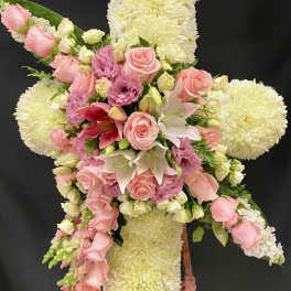 Standing floral cross of white mums with cascading pink roses, lilies, and lavender blooms on a wooden easel.