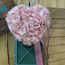 Heart-shaped arrangement of light pink roses with a pink ribbon on a green easel stand.