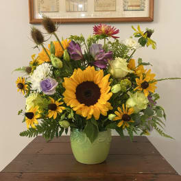 Mixed arrangement with sunflowers, yellow daisies, roses and purple blooms in a green ceramic vase