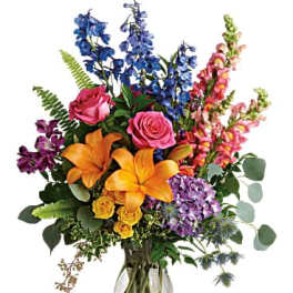 Colorful mixed bouquet in a clear glass vase with lilies, roses, and tall blue blooms