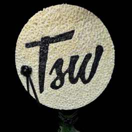 Round white floral tribute with black lettering and ribbon, mounted above a small floral base