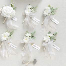 Set of white silk rose boutonnieres with ribbons and groomsmen tags on a light background