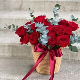 Red roses in a woven basket with a burgundy ribbon and eucalyptus
