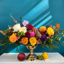 Colorful mixed flower arrangement in a gold vase with fruit on a white table