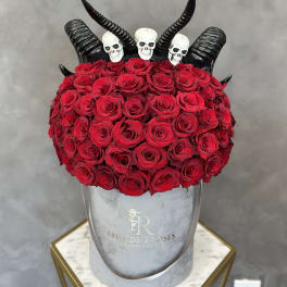 Red roses arranged in a gray hat box with black horn decorations and skull toppers